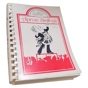 Puede incluir: Un libro de cocina encuadernado en espiral titulado "Apron Strings" con una cubierta roja y blanca. La cubierta presenta un chef de dibujos animados con un delantal con teclas de piano, dirigiendo. El texto "The Women's Committee of the Detroit Symphony Presents" también está en la cubierta.