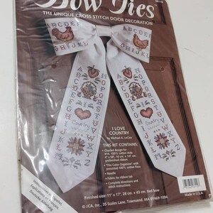 May include: A cross stitch kit for a door decoration called "Bow Ties" featuring a country theme. The kit includes a pre-finished ribbon, floss, needle, fabric for a ribbon tab, and instructions. The finished size is 11 inches by 17 inches.