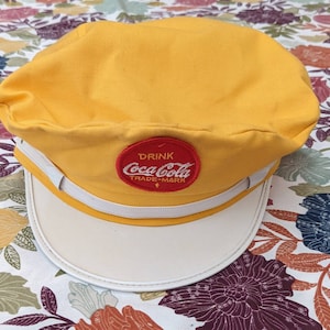 May include: A yellow and white vintage Coca-Cola cap with a red and white embroidered logo that says "Drink Coca-Cola Trade-Mark".