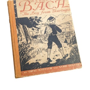 May include: A vintage book cover with a black and white illustration of a young boy walking on a path. The book title is "Sebastian Bach The Boy from Thuringia" and the author is "Opal Wheeler and Sybil Deucher".