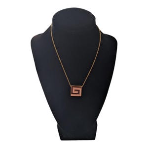 May include: A gold chain necklace with a square pendant featuring a geometric design. The pendant is a rose gold color.
