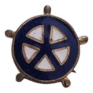 May include: A gold-colored pin with a blue and white enamel design. The design is a stylized wheel with five spokes. The pin is in the shape of a wheel with five spokes.