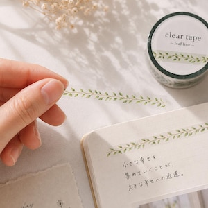 Puede incluir: Un rollo de cinta adhesiva transparente con la etiqueta clear tape - leaf line, que presenta un patrón repetitivo de hojas verdes. Una tira de cinta está extendida, mostrando el delicado diseño de follaje verde sobre una página con texto japonés.