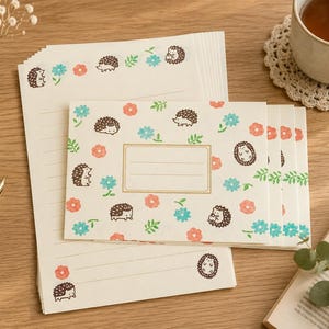 May include: Stationery set featuring a cream-colored paper with a repeating pattern of brown hedgehogs, blue and coral flowers. Includes lined paper and envelopes. A pen and a cup of tea are also visible.
