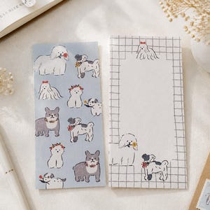 Puede incluir: Dos hojas de papel de carta rectangulares. La hoja izquierda es de color azul claro con un patrón repetitivo de varios perros ilustrados con coronas de flores. La hoja derecha es blanca con un borde de cuadrícula negra y perros.