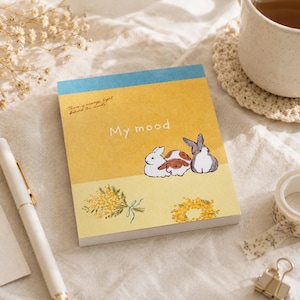 Könnte beinhalten: Ein rechteckiger Notizblock mit gelbem Einband, der drei illustrierte Kaninchen, einen Strauß gelber Blumen und einen Kranz zeigt. Der Text "My mood" und "There is always light behind the clouds" ist in weißer und brauner Schrift zu sehen.