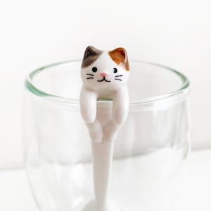 以下が含まれることがあります： 透明なグラスの縁にぶら下がっている、茶色、白、オレンジ色の頭を持つ白いセラミック製の猫のスプーン。猫のスプーンは笑顔で、カップやグラスの縁に置けるようにデザインされています。