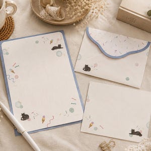 Peut inclure: Un ensemble de papeterie comprenant une feuille de papier à lettres, une enveloppe à rabat festonné et une carte de correspondance. Chaque élément est crème avec un motif de chats noirs, de glaces colorées et de petits cercles sur un bord bleu.