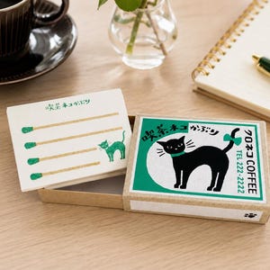 May include: A matchbox with a green and white label featuring a black cat silhouette with a green bow. The box is open, revealing matches with green tips and a green cat illustration. A cup of coffee and a notepad are in the background.