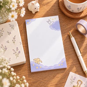May include: A rectangular notepad featuring a white center with light purple clouds and yellow stars at the top and bottom corners. A small white rabbit peeks from the top cloud, while a light brown rabbit rests in the bottom cloud.