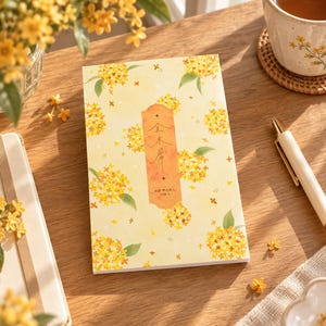 Peut inclure: Un bloc-notes rectangulaire au fond jaune pâle orné d'un motif répétitif de petites fleurs jaunes et de feuilles vertes. Une étiquette orange verticale au centre affiche du texte japonais et des petites croix, avec du texte en bas.