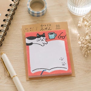 May include: A rectangular notepad with 30 sheets featuring a red border and a white center. The design includes a black and white cat, a blue mug, and black glasses. The top header displays Japanese text and the words 30 Sheets. The notepad is mounted on a brown cardboard backing.