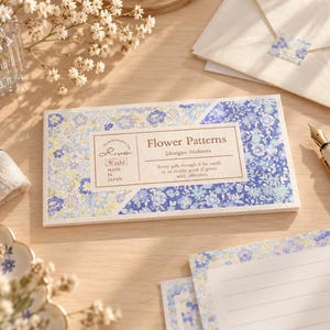 Könnte beinhalten: Eine rechteckige Packung Washi-Papierbögen mit blau-weißem Blumenmuster. Das Etikett trägt die Aufschrift Flower Patterns, 2designs 16sheets, Risette, Washi, MADE IN JAPAN und enthält einen kurzen Text über Geschenke und Zuneigung.