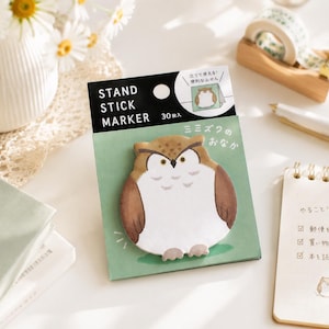 Könnte beinhalten: Eine Packung mit 30 eulenartigen Haftnotizen auf einer grünen Trägerkarte mit dem Text STAND STICK MARKER. Die Eule ist braun und weiß mit einem strengen Ausdruck. Die Verpackung enthält eine kleine kreisförmige Grafik, die die Notizen stehend zeigt.
