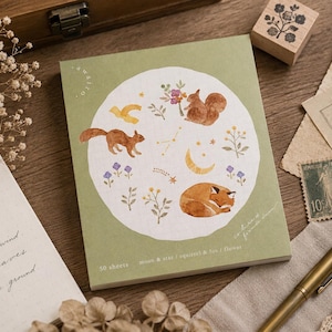 May include: A rectangular notepad with a sage green cover featuring a white circular illustration of two squirrels, a sleeping fox, a crescent moon, stars, and small floral sprigs. The text reads 50 sheets, moon & star, squirrel & fox, flower.
