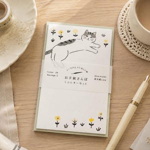 Könnte beinhalten: Ein Schreibwarenset bestehend aus einem weißen Papierblock mit einer grau-weißen Katzenillustration und gelben Blumen. Ein weißes Papierband umschließt die Mitte und ist mit japanischem Text, "Letter 20", "Envelope 5" und "MINO WASHI" bedruckt.