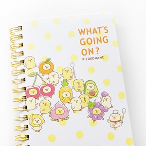 Pode incluir: Um caderno branco de espiral com bolinhas amarelas e uma ilustração colorida de pintinhos e frutas de desenho animado. A capa do caderno diz "What's Going On? Piyokomame".