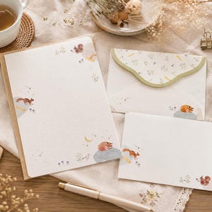Peut inclure: Un ensemble de papeterie comprenant une feuille de lettre rectangulaire, une enveloppe pliée avec un rabat festonné et une petite carte. Chaque élément est orné d'illustrations de petits écureuils bruns, de fleurs et d'un croissant de lune.