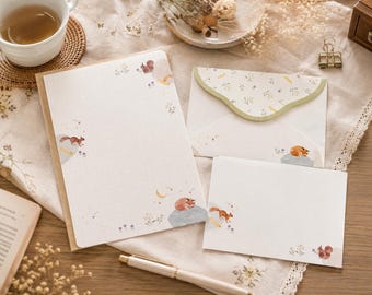 Puede incluir: Un juego de papelería que incluye una hoja de carta rectangular, un sobre plegado con solapa festoneada y una tarjeta pequeña. Cada pieza está decorada con ilustraciones de pequeñas ardillas marrones, flores y una luna creciente.