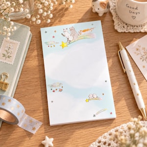 Può includere: Un blocco note rettangolare con uno sfondo color azzurro cielo, decorato con illustrazioni ad acquerello di gatti che cavalcano stelle cadenti, piccoli pianeti colorati e varie stelle. Il blocco ha un'ampia area centrale vuota.