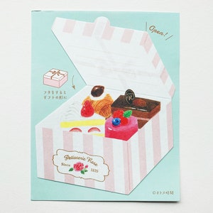 Puede incluir: Una caja de rayas rosas y blancas con una tapa que se abre para revelar una variedad de pasteles. La caja tiene una etiqueta que dice "Patisserie Rose" y "Since 1835". El texto "Open!" está escrito en la tapa de la caja.