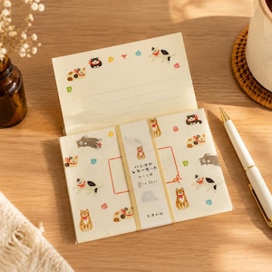 Pode incluir: Um conjunto de papelaria com papel de cor creme decorado com pequenas ilustrações de gatos usando coleiras vermelhas. O papel é mantido unido por uma faixa com o texto japonês "ハンコのレターセット ねことり". O conjunto inclui folhas pautadas.