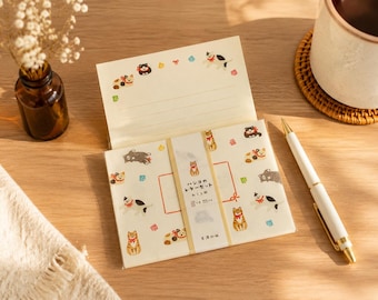 Puede incluir: Un juego de papelería con papel de color crema decorado con pequeñas ilustraciones de gatos con collares rojos. El papel se mantiene unido por una banda con el texto japonés "ハンコのレターセット ねことり". El conjunto incluye hojas con líneas.