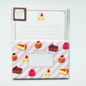 Op de afbeelding: Een set briefpapier met een roze-wit gestreepte envelop met verschillende kleurrijke gebakjes en taarten. De envelop heeft een wit label met de tekst "Patisserie Rose Since 1975". Het briefpapier heeft een witte achtergrond met een gouden rand en een kleine illustratie van een taart met een aardbei erop.