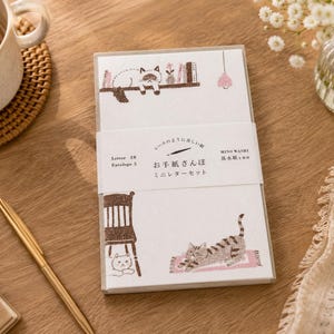 Könnte beinhalten: Ein Briefpapier-Set mit Illustrationen von Katzen und Möbeln. Das Set enthält Briefpapier und Umschläge mit japanischem Text auf der Verpackung. Die Illustrationen sind in Braun- und Grautönen gehalten, mit einer Katze, die auf einem Regal liegt, und einer anderen, die sich auf einem Teppich streckt.