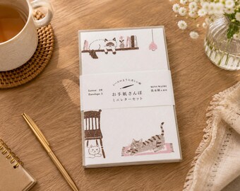 Könnte beinhalten: Ein Briefpapier-Set mit Illustrationen von Katzen und Möbeln. Das Set enthält Briefpapier und Umschläge mit japanischem Text auf der Verpackung. Die Illustrationen sind in Braun- und Grautönen gehalten, mit einer Katze, die auf einem Regal liegt, und einer anderen, die sich auf einem Teppich streckt.