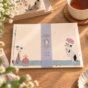 Op de afbeelding: Een crèmekleurige briefpapier set met een blauwe papieren band met de tekst "IOCORO COMUGI LETTER SET" en iconen voor 8 vellen en 4 enveloppen. Het papier toont een kat die een roze bloem vasthoudt en een vaas met blauwe bloemen.
