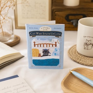 May include: A light blue rectangular sticky note pad featuring the title A Story of the Cat Who Stays Up Late. The cover illustration depicts a black cat in a bed with a brown teddy bear. The pad contains 40 sheets and has decorative gold filigree borders.