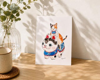 以下が含まれることがあります： 2匹の猫の水彩イラスト。1匹の猫がもう1匹の上に座っており、どちらも青い首輪をしています。下の猫は白と黒の模様で赤い帽子をかぶっています。上の猫はオレンジ、白、黒です。アートワークは白いカードにあります。