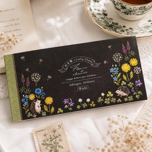 Può includere: Un blocco di carta washi rettangolare nero con illustrazioni in stile ricamo raffiguranti ricci, denti di leone gialli, fiordalisi blu e fiori viola. Il testo recita "Lucky Charm Fleurs et chance 2designs 16sheets Washi".