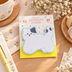 Könnte beinhalten: Ein Pack mit 20 katzenförmigen Haftnotizen mit der Aufschrift mofu mofu KATANUKI FUSEN. Die weißen Papiernotizen zeigen eine Illustration einer Katze mit grauen Ohren und blauem Umriss auf einer weißen Trägerkarte mit bunten Rauten.
