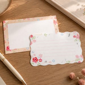 Puede incluir: Dos tarjetas de papelería rectangulares con bordes festoneados. Una tarjeta está en blanco con un borde floral, mientras que la otra presenta líneas horizontales, un patrón floral y pequeños iconos de notas musicales en rosa, rojo y azul.