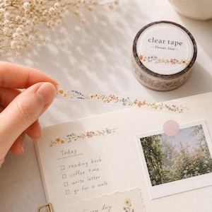 May include: A roll of clear tape labelled "clear tape - flower line -" featuring a delicate floral pattern. A strip of the tape is pulled out, displaying small, colourful flowers in shades of pink, blue, and yellow on a transparent background.