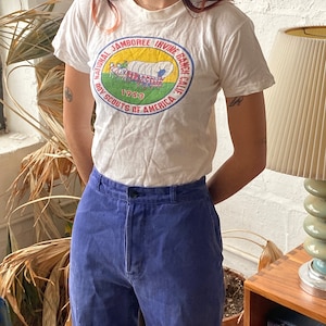 May include: White t-shirt with a vintage Boy Scouts of America National Jamboree Irvine Ranch, California 1953 patch. The patch features a wagon train with a red, white, and blue design.
