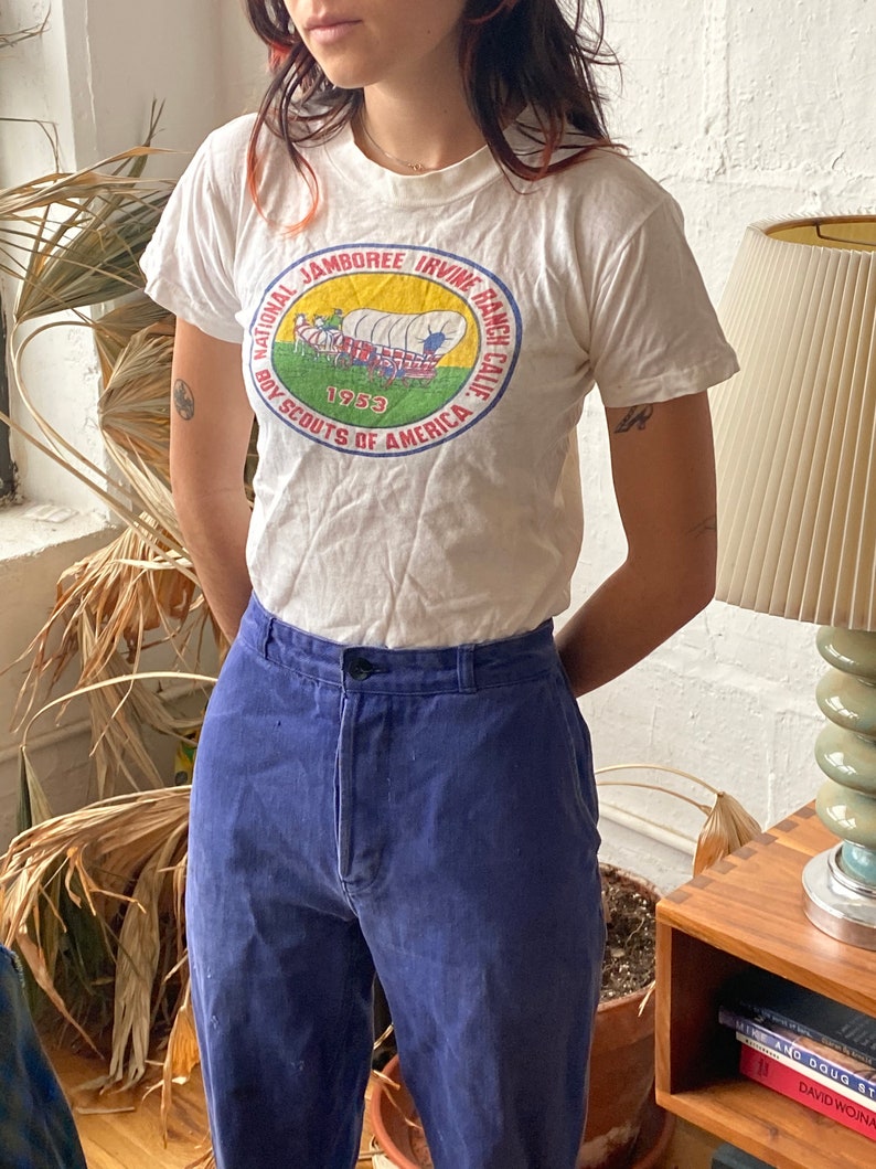 May include: White t-shirt with a vintage Boy Scouts of America National Jamboree Irvine Ranch, California 1953 patch. The patch features a wagon train with a red, white, and blue design.