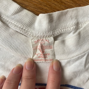 May include: White t-shirt with a red tag that says "Official Out" and "T" Shirt Small Chest Meas 28-30". The shirt has a blue and yellow stripe across the bottom with the word "Serving" in red.