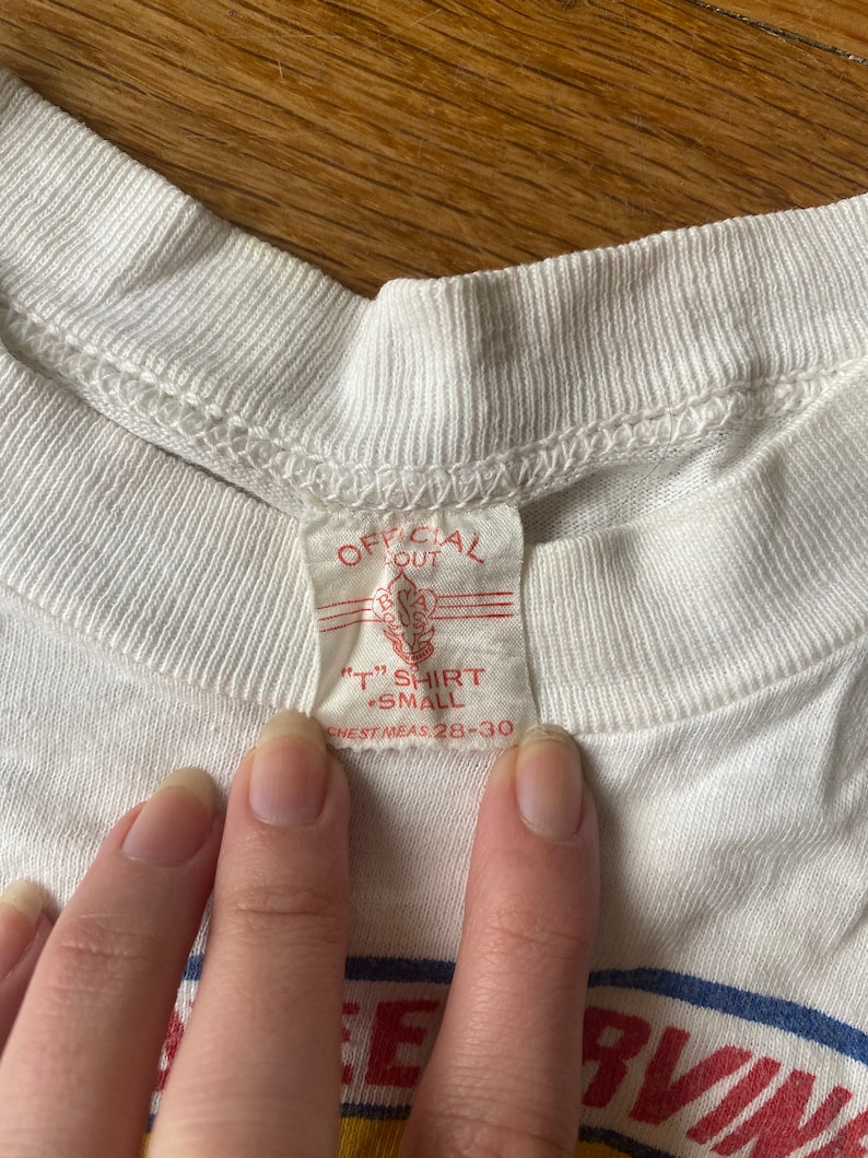 May include: White t-shirt with a red tag that says "Official Out" and "T" Shirt Small Chest Meas 28-30". The shirt has a blue and yellow stripe across the bottom with the word "Serving" in red.