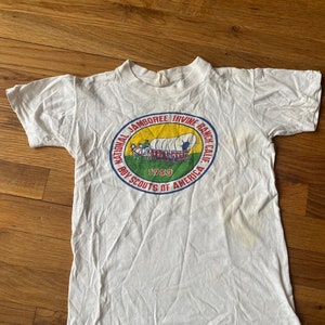 May include: White t-shirt with a circular graphic of a wagon train and the text "National Jamboree Irvine Ranch Calif. Boy Scouts of America 1953"