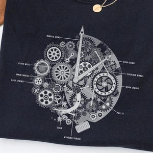 May include: Navy blue t-shirt with a detailed clock mechanism design in grey tones. The design includes labels like "Minute Hand," "Escape Wheel," and "Main Spring." A gold necklace is visible in the upper right corner.