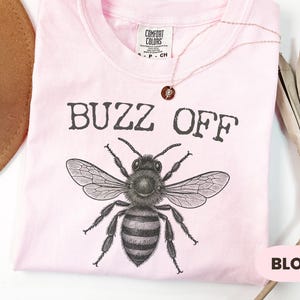 May include: A light pink t-shirt with the words "BUZZ OFF" above a detailed illustration of a bee. The shirt is folded, and a gold necklace with a pendant is draped across it. The word "BLOSSOM" is in a pink rectangle.