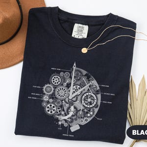 May include: Black crew neck t-shirt featuring a detailed grey clock mechanism graphic. The design includes gears, hands, and labeled components. A gold necklace and a brown hat are also in the image.