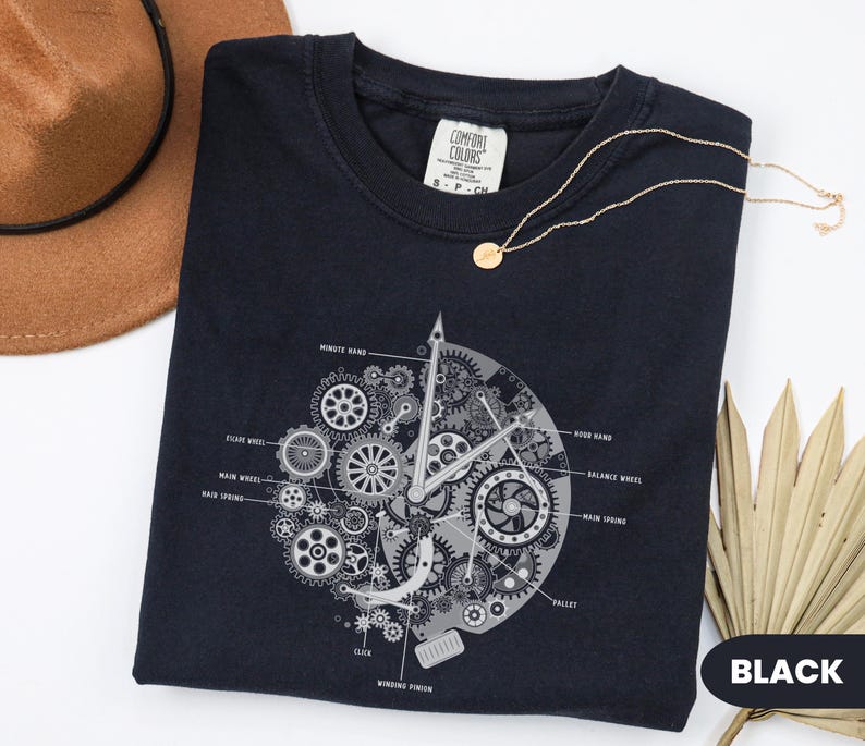May include: Black crew neck t-shirt featuring a detailed grey clock mechanism graphic. The design includes gears, hands, and labeled components. A gold necklace and a brown hat are also in the image.