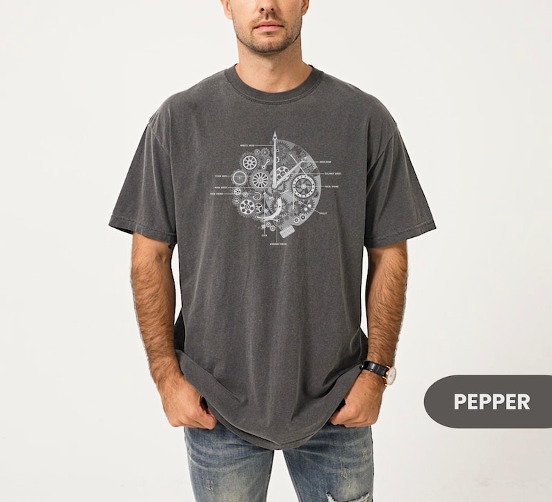 May include: A dark grey t-shirt with a white mechanical clock design. The design features gears and cogs. The shirt is made of a soft material and has a relaxed fit. The word "PEPPER" is in a grey rectangle. The shirt is a casual wear item.