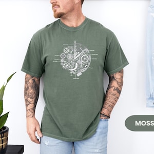 May include: A moss green t-shirt with a white graphic of clock gears and mechanisms. The design is centered on the shirt. The word "MOSS" is in a green rectangle in the bottom right corner.