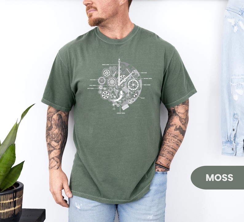 May include: A moss green t-shirt with a white graphic of clock gears and mechanisms. The design is centered on the shirt. The word "MOSS" is in a green rectangle in the bottom right corner.