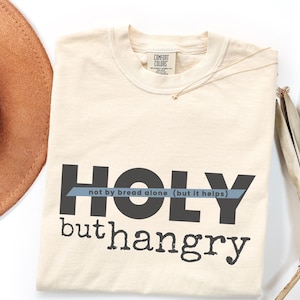 Op de afbeelding: Crèmekleurig T-shirt met de woorden "HOLY" en "hangry" in donkergrijze blokletters. De zin "not by bread alone (but it helps)" is gedrukt in een lichtblauwe banner over het woord "HOLY". Een bruine hoed en een gouden ketting zijn ook zichtbaar.